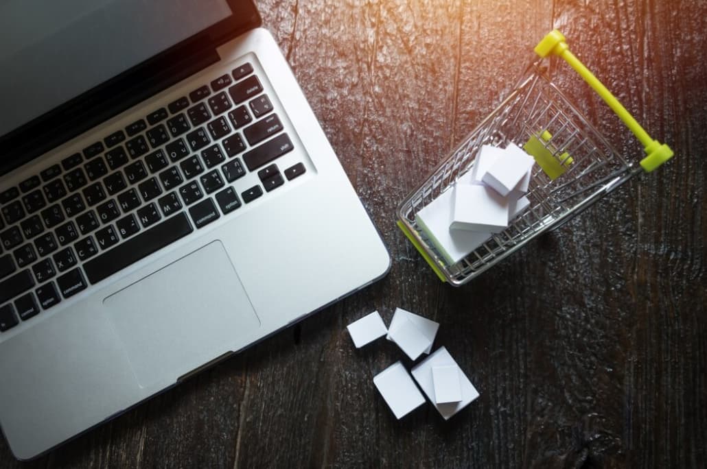 A laptop with a miniature shopping cart containing white boxes next to it